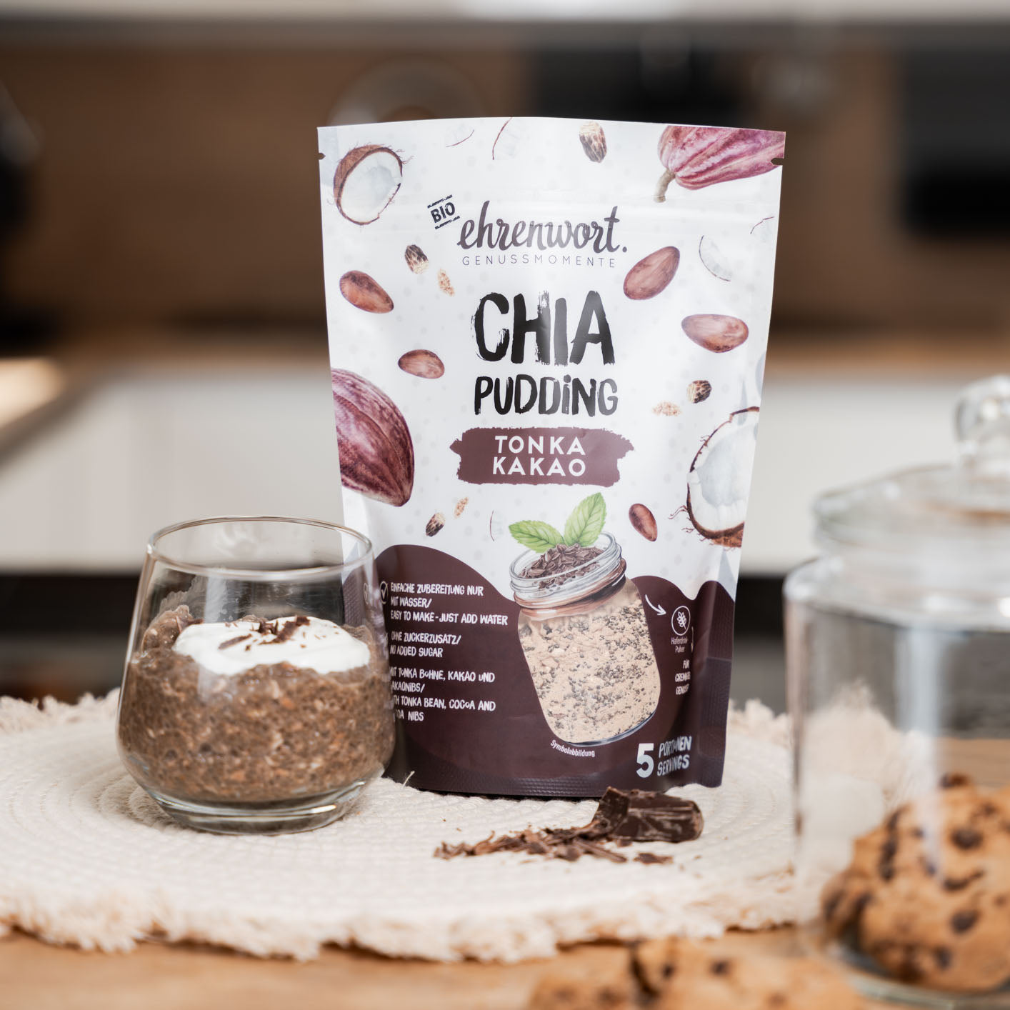 ehrenwort BIO Tonka Kakao Chia Pudding Verpackung neben zubereiteter Portion in einer Küche auf einem Stoffuntersetzer neben Keksen